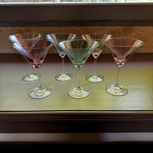 May include: Set of six clear glass martini glasses with iridescent rainbow coloured bases. The glasses are arranged in two rows of three on a wooden surface.
