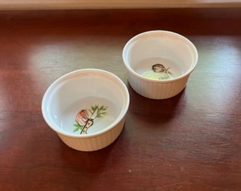 以下が含まれることがあります： 装飾的なデザインが施された白いセラミック製のラメイクインが2つ。1つのラメイクインには、緑の葉とナイフを持った赤いリンゴが描かれています。もう1つのラメイクインには、緑の葉と黄色の蝶が描かれています。