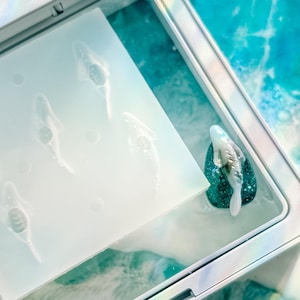 May include: A white ceramic mug with a black handle and the text "But First, Coffee" in a bold, black font. The mug is set against a blurred background, suggesting a cozy setting. The mug appears to be approximately 4 inches tall.