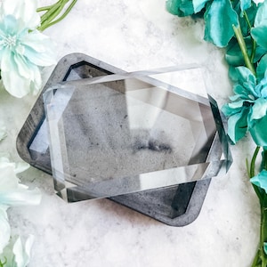 May include: A rectangular tray with a dark grey, textured finish and a clear, faceted, geometric glass cover resting on top. The tray features bevelled edges and a smooth interior surface, while the transparent lid displays sharp, angular lines.