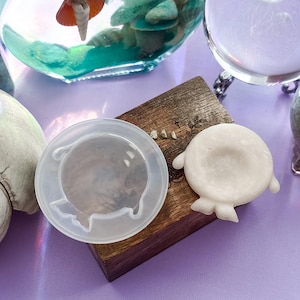 Puede incluir: Un molde de silicona circular y translúcido con un diseño de pequeña criatura estilizada, colocado junto a una pieza blanca opaca de la misma forma, ambos sobre un bloque de madera rectangular con un acabado de veta natural.