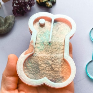 Puede incluir: Un molde de resina con forma de hueso, con un borde blanco y una mezcla de colores naranja, dorado y verde en el interior. Una pequeña clavija blanca está en la parte superior. Otro molde similar es visible en el fondo.