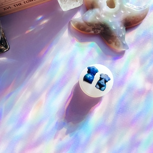 May include: A white, round object with two blue torso illustrations. The image is set against a backdrop of iridescent light and includes a crystal starfish and a tarot card.