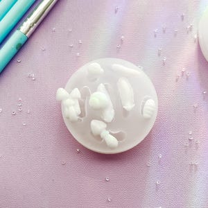 May include: A white silicone mould featuring several small, mushroom-shaped designs. The mould sits on a light purple surface, with a few light blue paint brushes and small, clear droplets scattered around. Another similar mould is visible.
