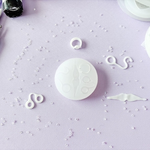 Puede incluir: Una colección de suministros de artesanía de resina blanca, que incluye un molde redondo con diseño de luna, varios dijes y pequeñas cuentas, dispuestos sobre una superficie de color lila claro. También se ven varios pinceles con mango azul claro.