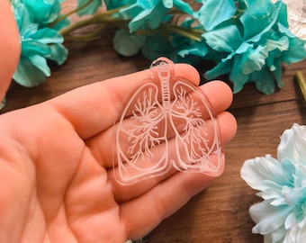May include: A transparent acrylic charm shaped like human lungs, featuring etched details of the bronchial tree. The charm is held in a hand, with a wooden surface and teal flowers in the background.