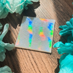 May include: A square, translucent coaster featuring a fine silver glitter finish and a holographic effect that displays vibrant, shifting rainbow colors across its surface when light hits the material at different angles.