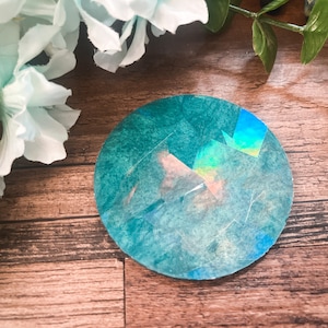 Könnte beinhalten: Ein runder Aufkleber mit einem tiefen blau-türkisfarbenen Aquarellhintergrund und einer reflektierenden holografischen Beschichtung, die geometrische Lichtmuster auf der Oberfläche erzeugt und eine glatte, kreisförmige Form aufweist.