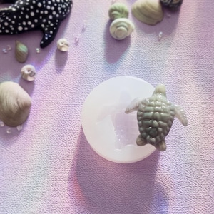 May include: A circular, translucent silicone mold featuring a sea turtle cavity, accompanied by a small, grey, textured sea turtle figurine resting on top of the mold, surrounded by various small seashells and a black and white spotted whale.