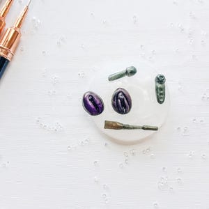 May include: An overhead shot shows a white surface with various small objects. Two purple, oval-shaped items are next to several green and bronze-colored pieces. Paintbrushes with black and gold handles are in the upper left corner, and small clear beads are scattered around.