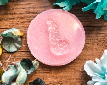 May include: A round, light pink resin piece with a white, textured letter "L" design. The resin is set against a wooden background, with floral accents in shades of blue, green, and yellow.