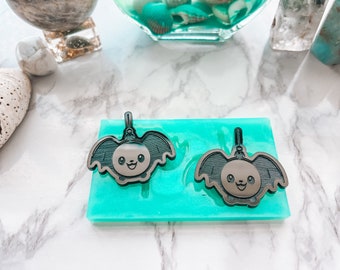 Puede incluir: Dos dijes en forma de murciélago de color gris oscuro con caras sonrientes y pequeños bucles en la parte superior, descansando sobre un molde de silicona rectangular de color turquesa brillante. Los dijes tienen texturas de alas.