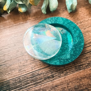 Puede incluir: Un posavasos redondo, transparente e iridiscente se encuentra sobre un posavasos texturizado de color verde azulado. Los posavasos están sobre una superficie de madera, con follaje verde en el fondo. El posavasos transparente tiene un diseño geométrico.