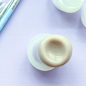 Pode incluir: Um molde de silicone bege, circular e côncavo, está posicionado ao lado de uma base circular branca. Três pincéis de maquilhagem azul-claros com pontas de silicone brancas estão dispostos na diagonal, junto a dois pequenos recipientes brancos.