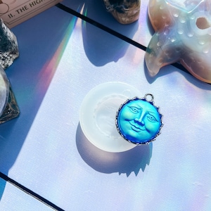 May include: A circular silicone mould featuring a recessed sun face design, paired with a vibrant blue sun-shaped pendant with a metallic silver-toned edge. The pendant displays a detailed facial expression and is positioned directly atop the mould.