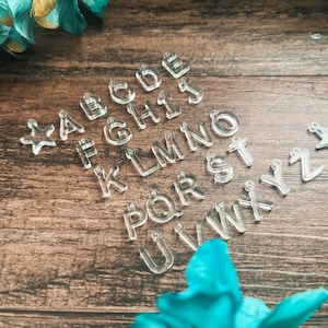 May include: A collection of clear acrylic alphabet letter charms from A to Z, each featuring a small loop at the top for attachment. The set includes two star-shaped charms, one positioned at the far left and one at the far right of the letters.