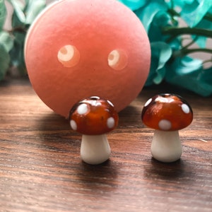 Puede incluir: Dos figuras de hongos de vidrio en miniatura con tallos blancos y tapas de color marrón rojizo con puntos blancos. Un objeto redondo grande de color melocotón con dos agujeros está detrás de los hongos. Los artículos están sobre una superficie de madera.