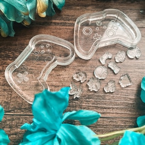 May include: Clear silicone moulds in the shape of a video game controller and various small shapes. The controller moulds have button details. The small shapes include a controller, a rose, and a butterfly. The items are on a wooden surface.