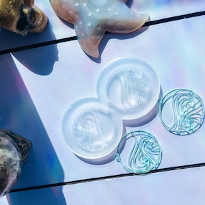 May include: A collection of clear resin molds and finished pieces with a wave-like design. The molds are in a connected circular shape, while the finished pieces are a translucent teal color. Other objects include a starfish and a tarot card.