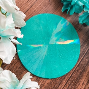 May include: A circular teal coaster featuring a reflective holographic strip across the center that catches the light. The surface has a smooth, flat finish with subtle radial lines radiating from the middle point of the disc.