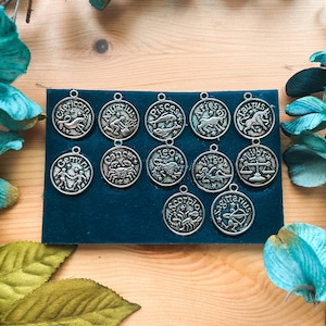 May include: A collection of silver-colored zodiac sign charms arranged on a dark teal felt surface. Each charm features a different astrological symbol and name, including Capricorn, Aquarius, and Pisces. The background includes wooden planks and teal floral accents.