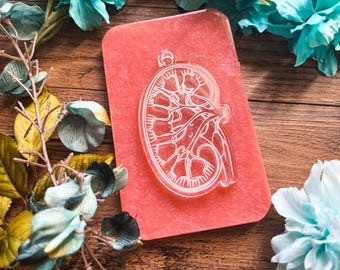 May include: A clear, kidney-shaped pendant with internal vein details, set on a textured, rectangular, coral-colored background. The pendant is suspended from a small loop. The image is decorated with teal and white flowers and foliage, set against a wooden surface.
