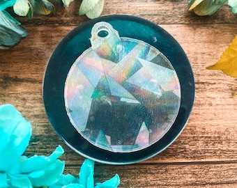 May include: A clear, round ornament with a geometric design, resting on a dark teal coaster. The ornament has a hole at the top for hanging and reflects a rainbow of colors. The background is a wooden surface with floral accents.