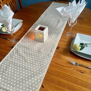 Könnte beinhalten: Ein beigefarbener Tischläufer mit weißem Herzmuster und gehäkeltem Rand liegt auf einem Holztisch. Ein weißer Kerzenhalter mit Herz-Ausschnitten und einer brennenden Kerze befindet sich auf dem Läufer. Weiße Servietten, Teller und Weingläser sind ebenfalls auf dem Tisch.