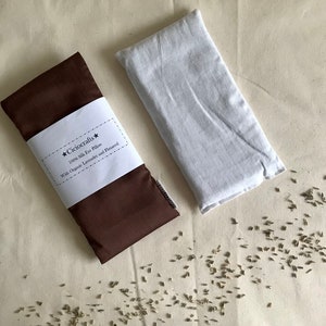 May include: A brown eye pillow with a white label that reads "Cicrocrafts* 100% Silk Eye Pillow With Organic Lavender and Flaxseed" and a white eye pillow. Scattered lavender seeds are visible on the surface.