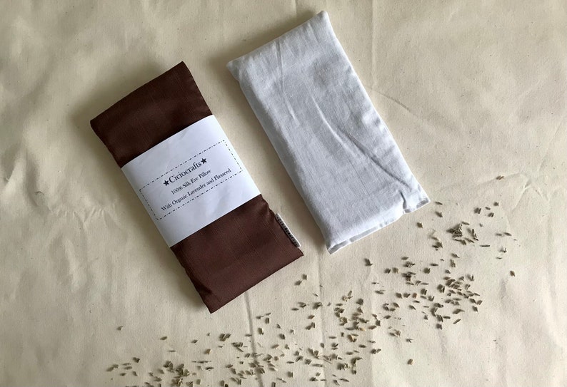 May include: A brown eye pillow with a white label that reads "Cicrocrafts* 100% Silk Eye Pillow With Organic Lavender and Flaxseed" and a white eye pillow. Scattered lavender seeds are visible on the surface.