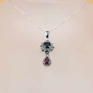 Puede incluir: Un collar de plata con un colgante que presenta una piedra cuadrada azul oscuro rodeada de pequeñas piedras transparentes. Debajo, una piedra roja en forma de lágrima también está rodeada de piedras transparentes. El colgante cuelga de una delicada cadena de plata.