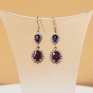 May include: A pair of drop earrings featuring a pear-shaped purple amethyst stone at the top and a larger oval-shaped deep red ruby stone at the bottom. Both stones are surrounded by a halo of small clear white stones set in a metallic frame.