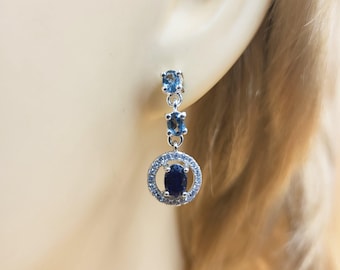 Puede incluir: Pendientes colgantes de plata con tres piedras preciosas azules. Las piedras superior y central son ovaladas, mientras que la inferior está engastada en un halo de piedras más pequeñas. Los pendientes se muestran en una oreja.