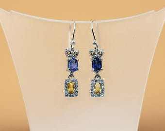Puede incluir: Un par de pendientes de plata con diseño de corona. Cada pendiente presenta una gema rectangular azul, una gema rectangular amarilla y pequeñas gemas transparentes. Los pendientes se exhiben sobre un soporte blanco sobre un fondo naranja claro.
