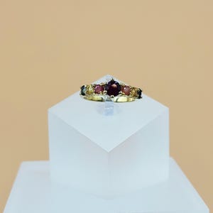 May include: A gold ring featuring a large, round, deep red gemstone at the center, flanked by smaller, colorful gemstones. The ring is set on a white, geometric display against a neutral background.