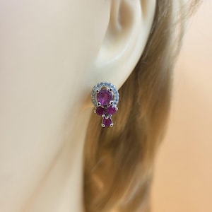 May include: A silver stud earring with pink gemstones. The earring has a large, round pink gemstone encircled by a halo of smaller, clear stones. Beneath the main stone, there is a cluster of three smaller pink gemstones.