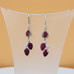 May include: Silver-tone drop earrings with three deep red, pear-shaped gemstones and a smaller, teardrop-shaped gemstone at the top, surrounded by clear stones. The earrings are displayed on a neutral-coloured stand. The earrings are displayed on a neutral-coloured stand.