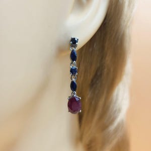 May include: Dangling earring with a silver setting. The earring features a series of dark blue gemstones and a large, oval-shaped, deep red gemstone at the bottom. The earring is shown on a person's ear.