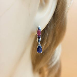 Può includere: Orecchini pendenti color argento con tre pietre preziose ovali. La gemma superiore è rosa intenso, quella centrale è viola scuro e quella inferiore è blu vibrante. Gli orecchini sono esposti su uno sfondo sfocato.