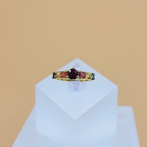 Peut inclure: Une bague en or ornée d'une grosse pierre rouge foncé au centre, flanquée de petites pierres colorées roses, oranges, jaunes et bleues. La bague est présentée sur un socle blanc géométrique, sur fond pêche.