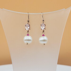 May include: Rose gold earrings with a light pink oval gemstone, a small red bead, and a large white pearl. The earrings hang from a gold hook. The background is a light peach color.