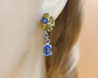Puede incluir: Un pendiente de plata con un diseño floral con piedras preciosas amarillas y azules. El pendiente tiene una gema azul colgante en forma de óvalo. El pendiente se lleva en la oreja de una persona.