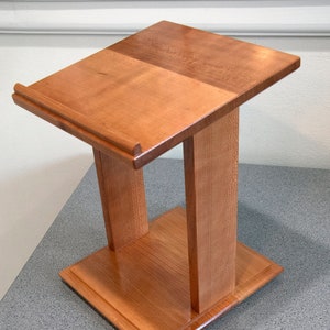 May include: A wooden lectern with a slanted top and a shelf underneath. The lectern is made of light brown wood and has a smooth finish.