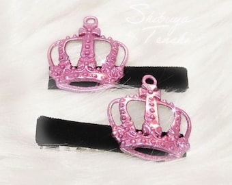 May include: Two pink crown hair clips with a sparkling finish, each attached to a black barrette. The crowns have intricate details and are set against a fluffy white background.
