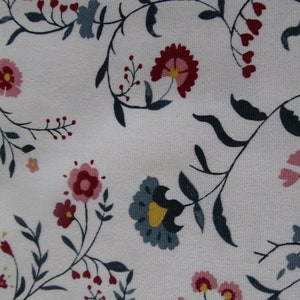 Op de afbeelding: Close-up van witte stof met een bloemenpatroon. Het ontwerp toont gestileerde bloemen in roze, rood en blauw, met donkergroene bladeren en stelen. De bloemen zijn verspreid over de stof.