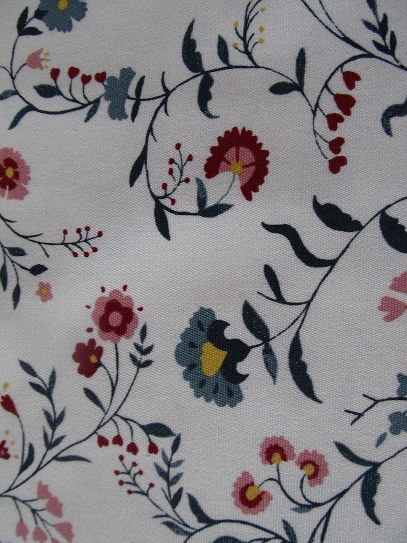 Op de afbeelding: Close-up van witte stof met een bloemenpatroon. Het ontwerp toont gestileerde bloemen in roze, rood en blauw, met donkergroene bladeren en stelen. De bloemen zijn verspreid over de stof.