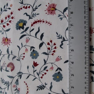 Op de afbeelding: Witte stof met een bloemenpatroon. Het ontwerp bevat roze, blauwe en rode bloemen met groene bladeren en stelen. Een liniaal is verticaal geplaatst, met metingen in centimeters en millimeters.