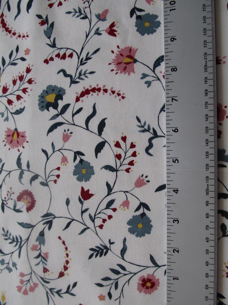 Op de afbeelding: Witte stof met een bloemenpatroon. Het ontwerp bevat roze, blauwe en rode bloemen met groene bladeren en stelen. Een liniaal is verticaal geplaatst, met metingen in centimeters en millimeters.