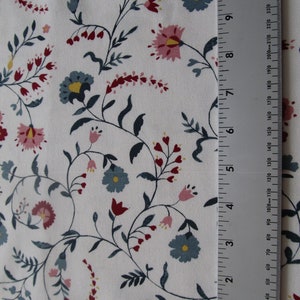 Op de afbeelding: Witte stof met een bloemenpatroon van roze, blauwe en rode bloemen en donkergroene ranken. Een liniaal is verticaal geplaatst en toont de afmetingen in centimeters.