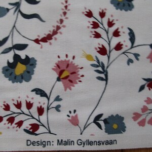 Op de afbeelding: Witte stof met een bloemenpatroon. Het patroon bevat roze, rode, blauwe en gele bloemen en groene bladeren. De tekst "Design: Malin Gyllensvaan" is zichtbaar.